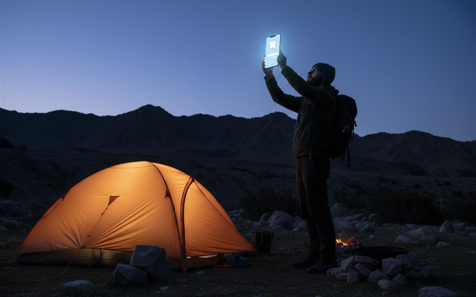 A camper by a tent at dusk holds up a phone showing a satellite icon as a message finally sends over the darkening ridge